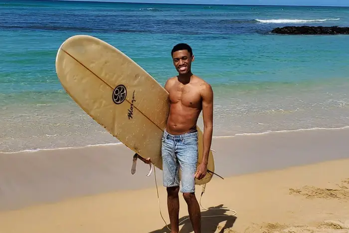 Tarique Peters stands on the beach, wearing jean shorts only, and holding a surfboard